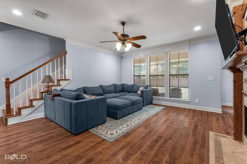 Wood-finish flooring, fireplace with stone surround, crown molding, staircase with stained wood handrail, and recessed lighting