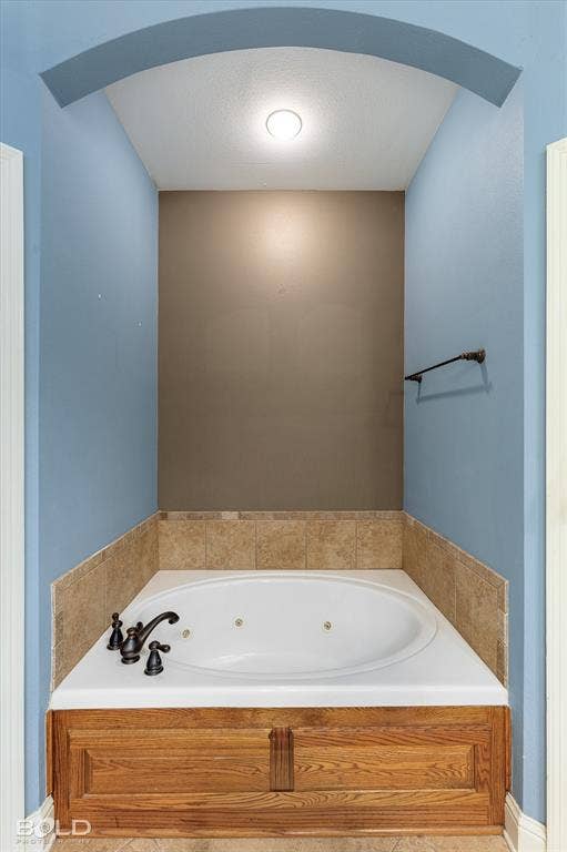 Jetted soaking tub featuring an arched recessed alcove, stone-finish tile surround, dark metal faucet hardware, and wood-finish cabinetry