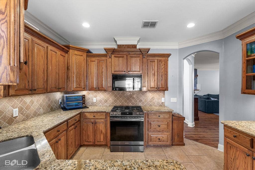Kitchen featuring custom wood cabinetry, natural stone countertops, a tile backsplash arranged in a diagonal pattern, stainless steel appliances, and crown molding