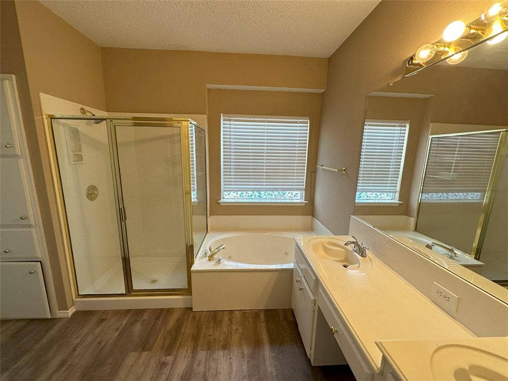 7800 Crestview Drive, Plano, TX 75024 White double vanity with integrated sinks, large soaking tub, separate glass-enclosed shower, wood-finish flooring, and built-in storage cabinetry