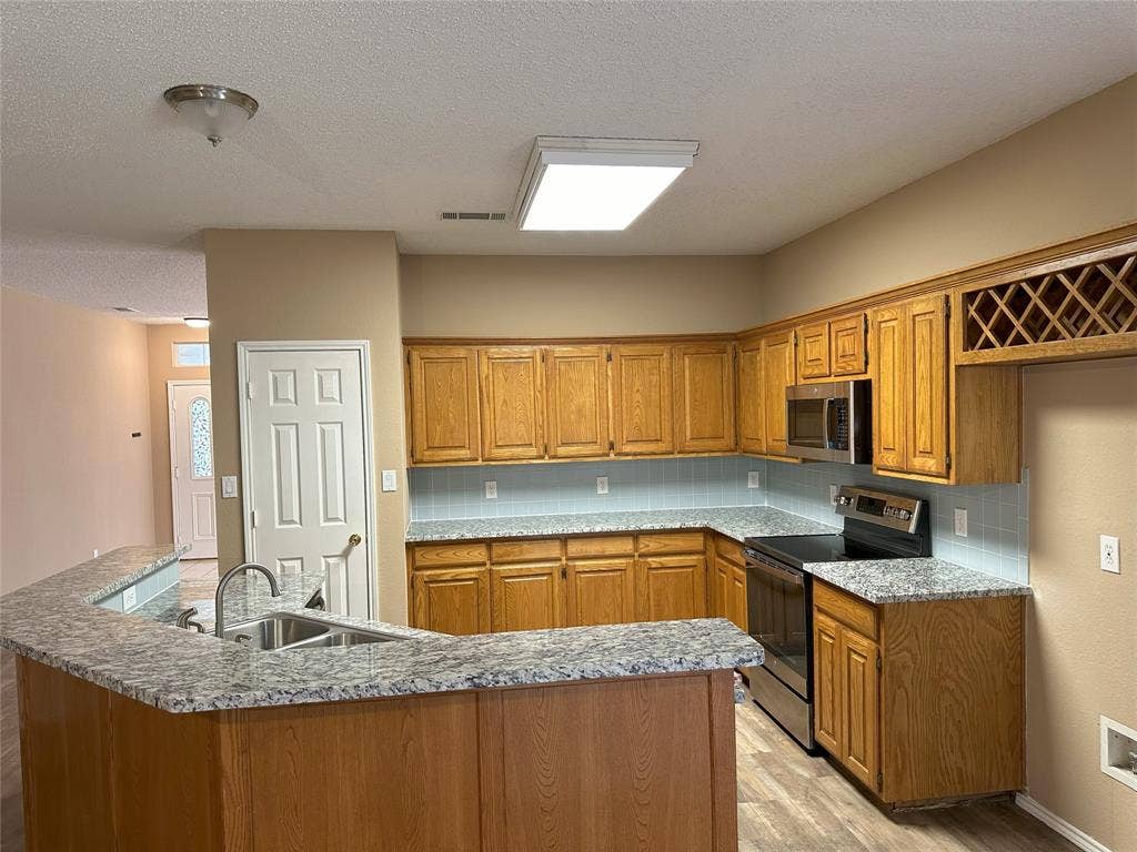 7800 Crestview Drive, Plano, TX 75024 Kitchen featuring a curved granite-finish breakfast bar, stainless steel appliances, light wood cabinetry, a tile backsplash, and wood-finish flooring