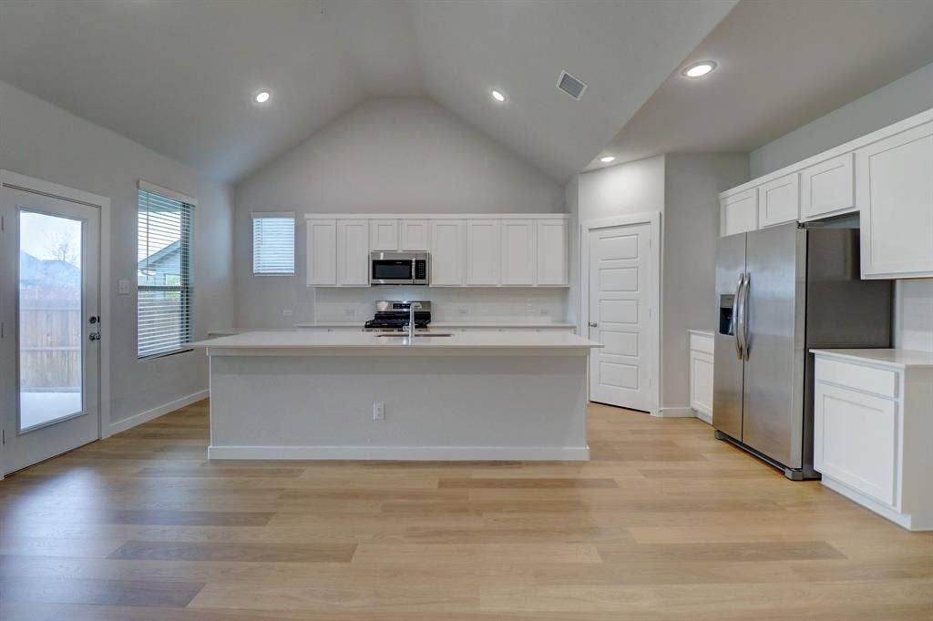 Vaulted ceiling with recessed lighting, wood-finish flooring, white cabinetry, central island with sink, and stainless steel appliances