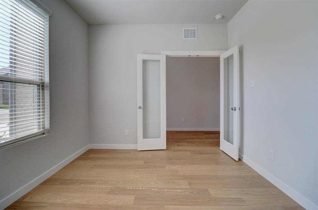 Light wood-finish flooring, white walls, double French doors with frosted glass, and a large window with blinds