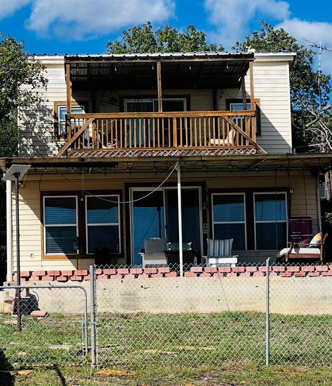 Two-story property with horizontal siding, two balconies with wood railings, a metal roof covering the upper balcony, and a chain-link fence