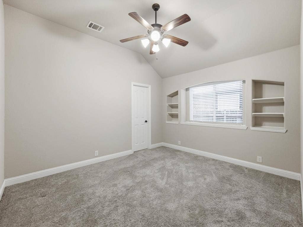 Vaulted ceilings featuring a central ceiling fan with multi-blade design, carpeted flooring, and arched built-in shelving flanking a window with integrated blinds