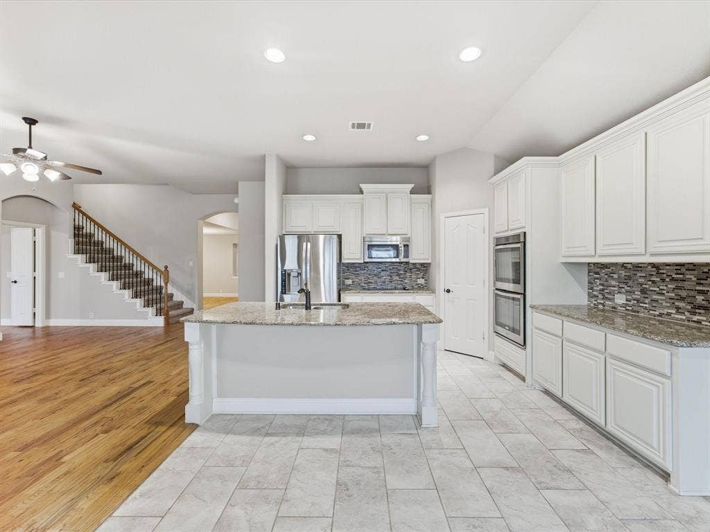 Kitchen featuring white shaker-style cabinetry, stone countertops, stainless steel double wall ovens, a multi-tone tile backsplash, and a central island with an integrated sink