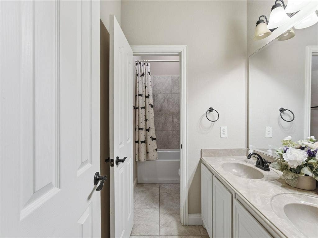 Dual-sink vanity with stone-surfaced countertop, matte black fixtures, full-width vanity mirror, ceramic tile flooring, and a bathtub with tile surround
