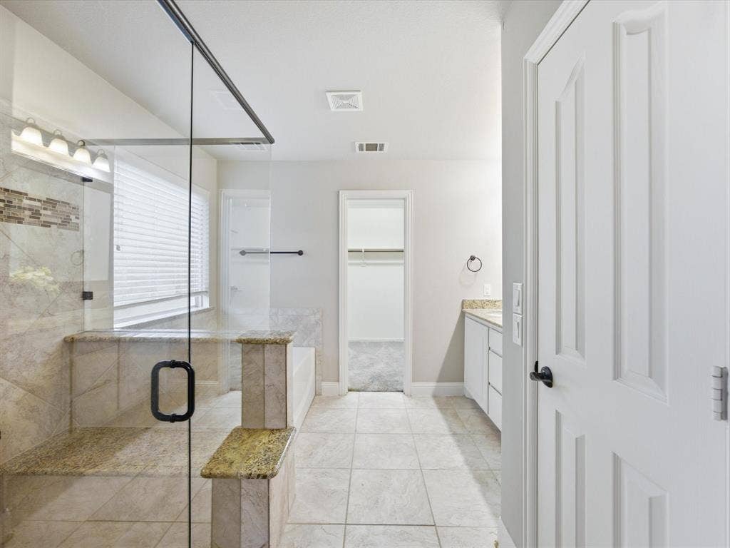 Walk-in shower with a glass enclosure, stone-finish tile surround, built-in bench seating, light-toned floor tile, and white cabinetry