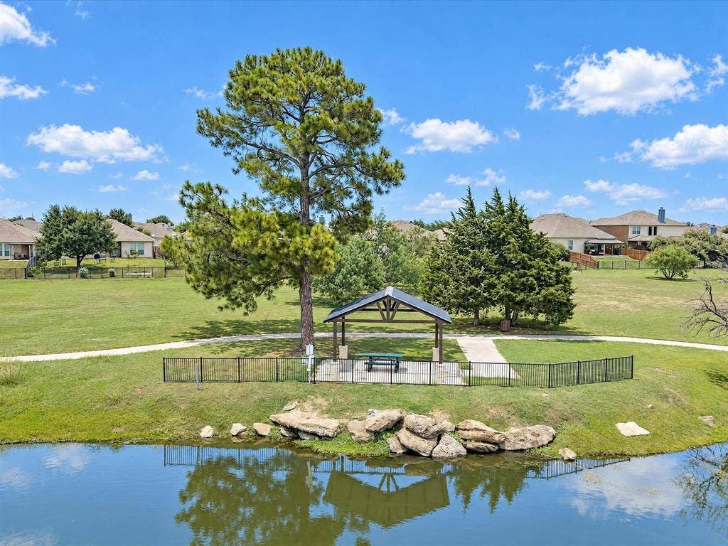 Open-air pavilion featuring a gabled metal roof and stone-clad pillars, situated along a pond edge with natural stone retaining wall accents, mature shade trees, and paved walking paths