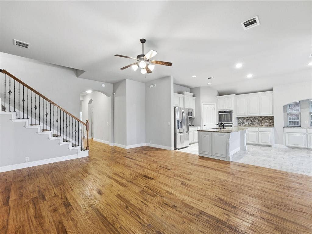 Open-concept area featuring wood-finish flooring, a staircase with decorative wrought-iron balusters, recessed lighting, and a kitchen equipped with white cabinetry, a center island, and stone-finish countertops