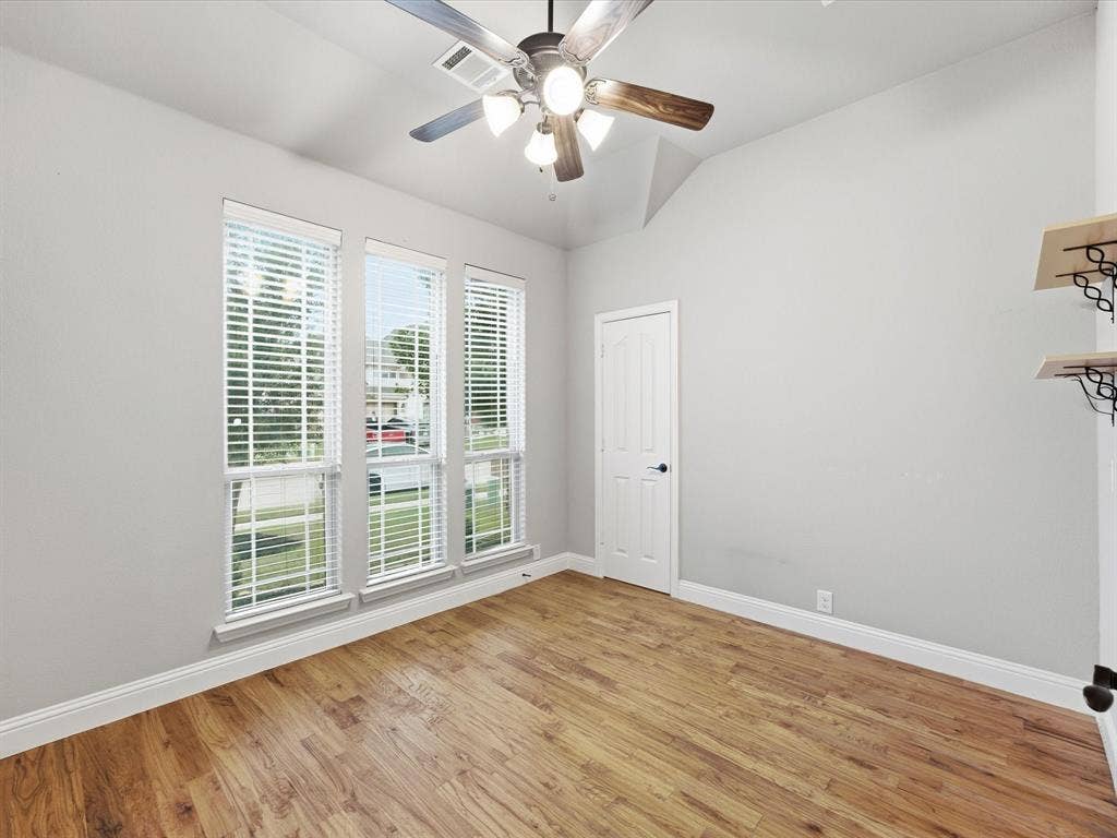 Room featuring wood-finish flooring, vaulted ceilings, three tall windows, a multi-light ceiling fan, and built-in wall shelving