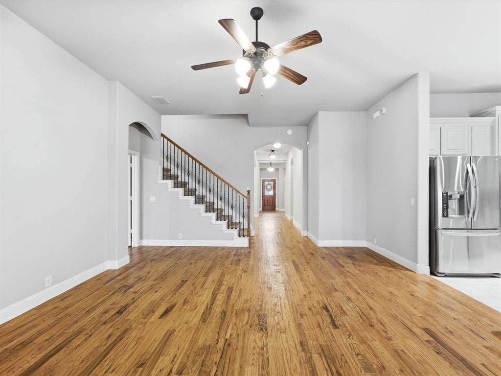 Open-concept interior featuring wood-finish flooring, a central ceiling fan with multi-light fixture, staircase with dark metal balusters and wooden handrail, and arched architectural entryways