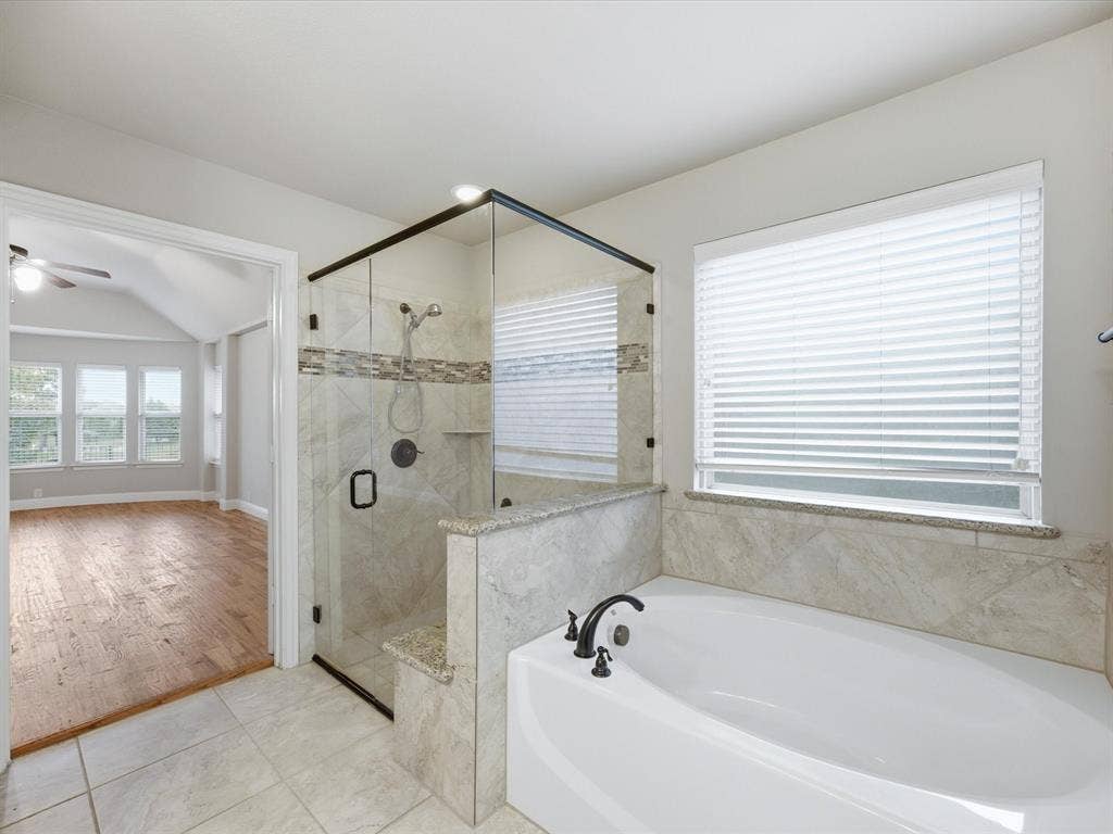 Soaking tub with stone-finish surround, glass-enclosed shower with decorative tile inlay, and large-format tile flooring