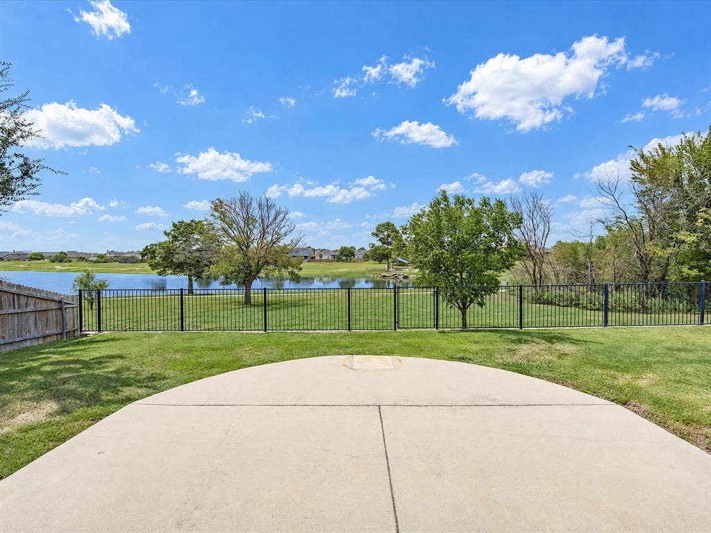 Concrete patio featuring a sodded lawn, black metal perimeter fencing, and mature trees