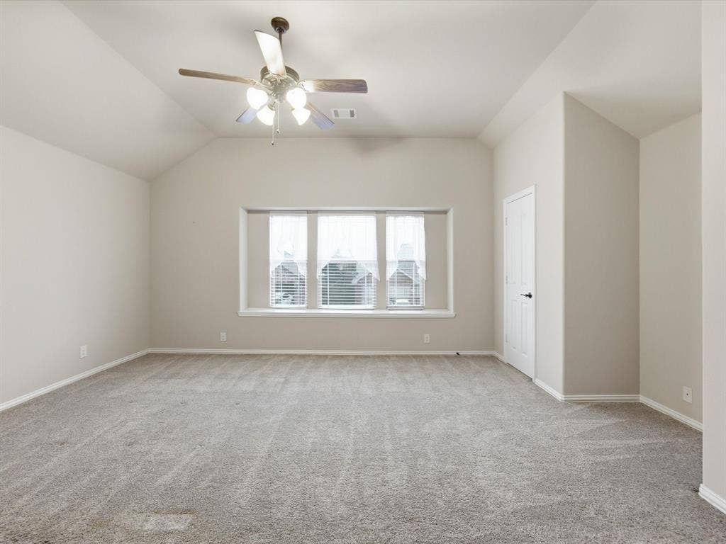 Vaulted ceiling, expansive bay window, ceiling-mounted fan with integrated lighting, neutral wall palette, and wall-to-wall carpeting