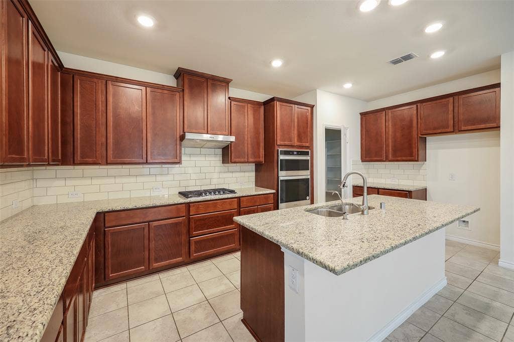 3681 Keechi Creek Drive, Celina, TX 75078 Kitchen featuring rich wood cabinetry, stone-surface countertops, subway tile backsplash, stainless steel double-wall ovens, and a central island with an integrated sink