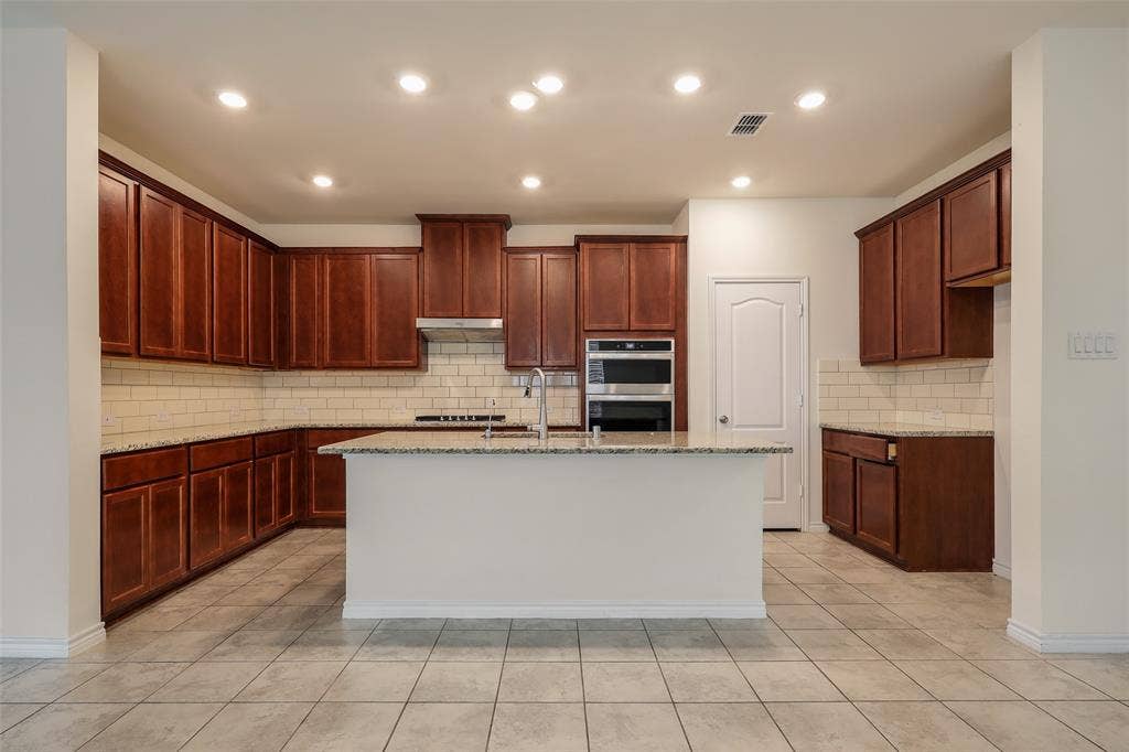 3681 Keechi Creek Drive, Celina, TX 75078 Kitchen featuring stained wood cabinetry, stone-surface countertops, subway tile backsplash, center island with sink, and stainless steel double wall ovens