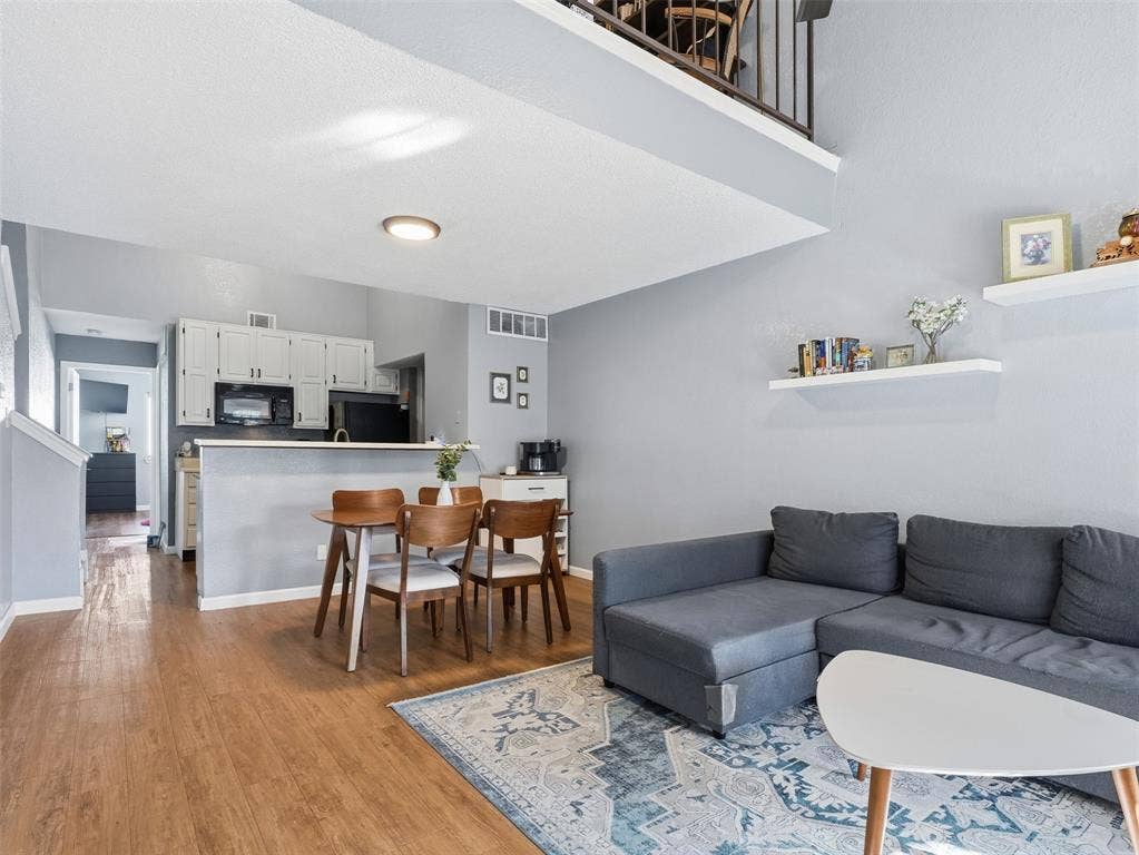 Open-concept area featuring wood-finish flooring, a lofted mezzanine with dark metal railing, white wall-mounted shelving, and a kitchen peninsula