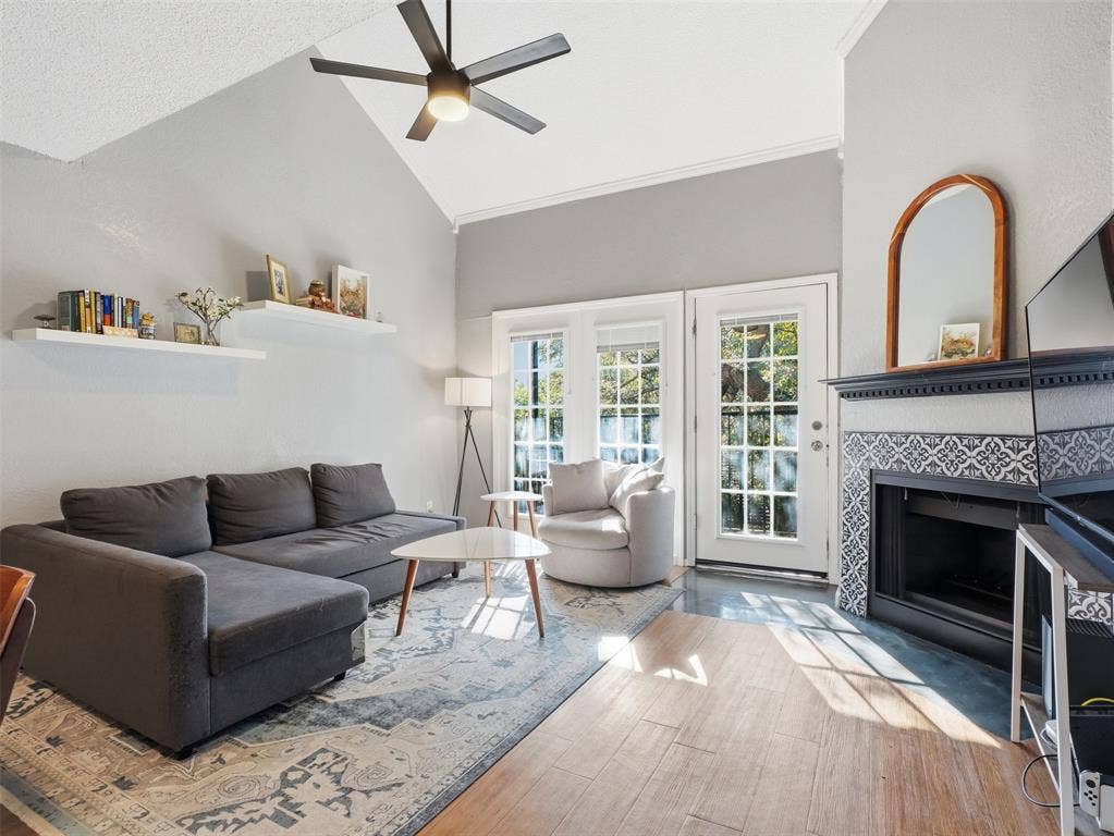 Vaulted ceilings featuring a central overhead fan, fireplace with patterned tile surround and a wood mantel, wood-finish flooring, built-in wall shelving, and grid-pane glass doors