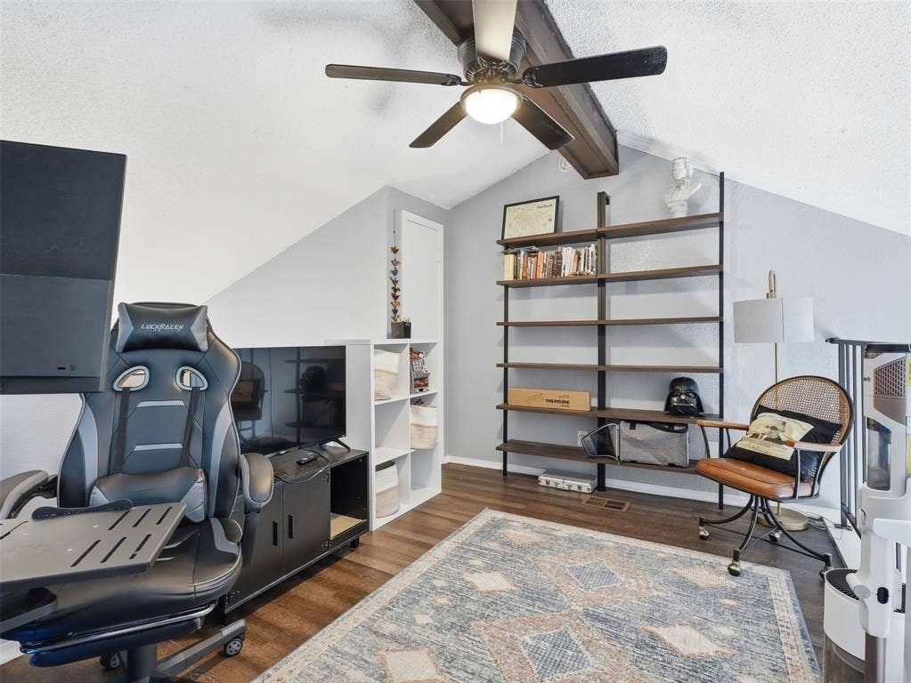 Sloped ceiling accented with a structural wood-finish beam, ceiling fan with integrated lighting, wall-mounted industrial-style shelving, and wood-finish flooring
