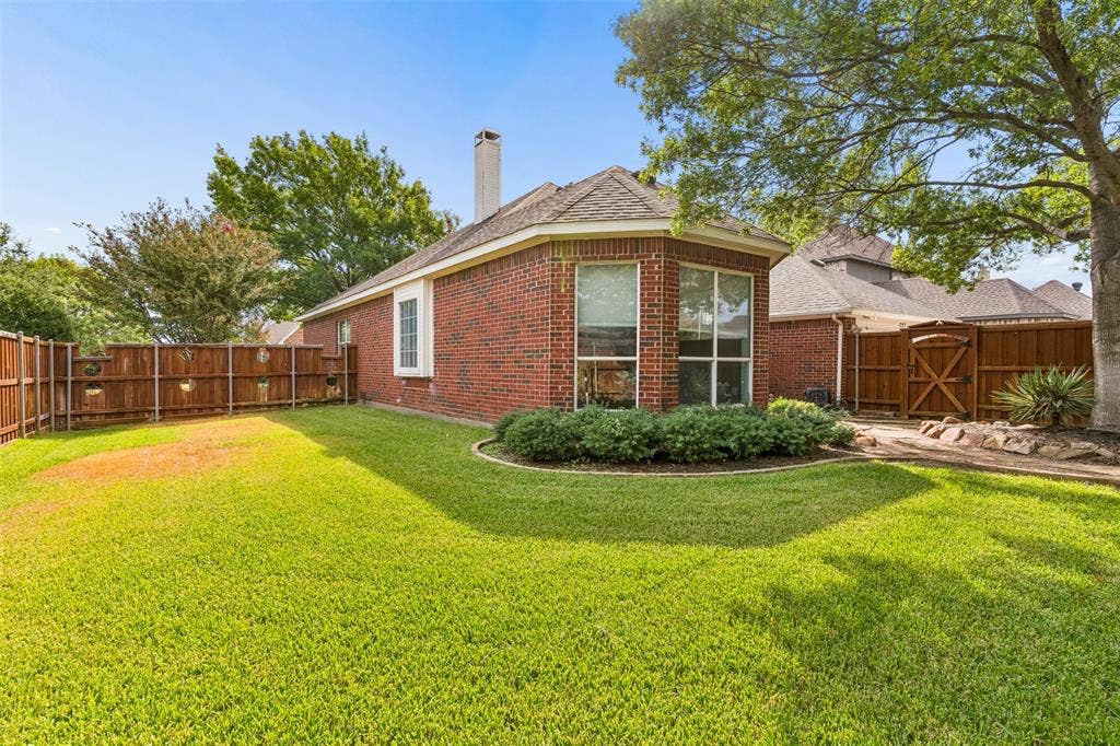 327 Hampton Court, Coppell, TX 75019 Brick exterior featuring a multi-faceted roofline, chimney, expansive lawn, mature trees, and wooden perimeter fencing
