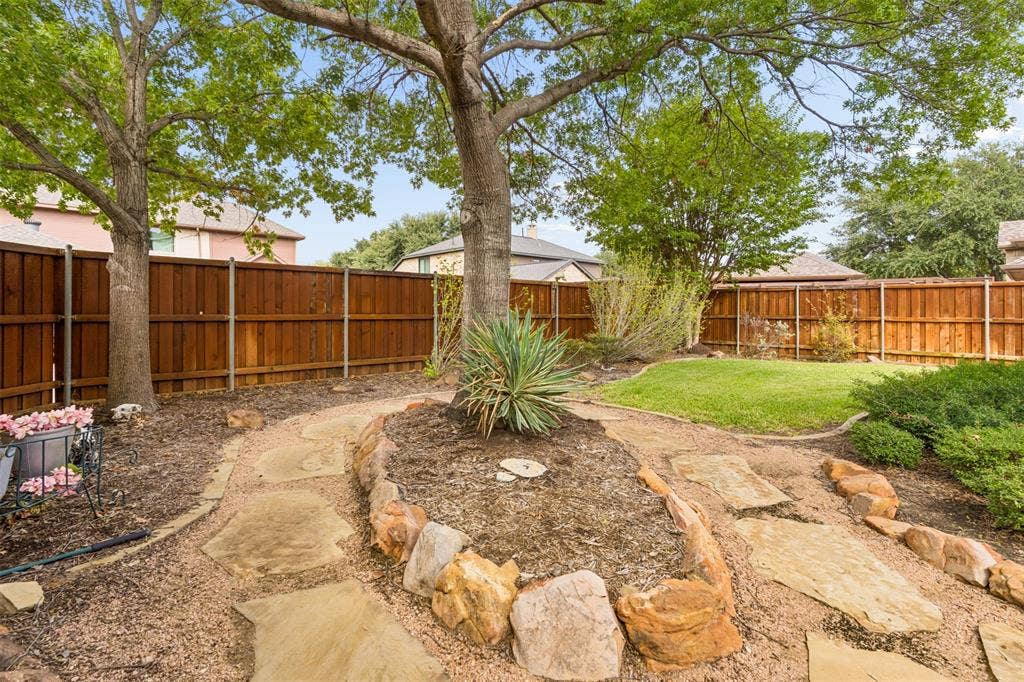 327 Hampton Court, Coppell, TX 75019 Backyard featuring a wood privacy fence, stone paver pathway, elevated stone-bordered garden bed, and mature trees