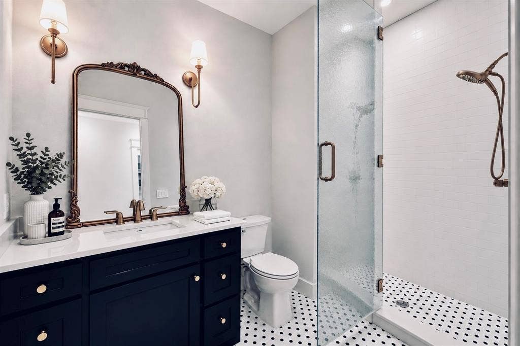 6651 Pinehurst Drive, Granbury, TX 76049 Dark vanity with integrated sink and brass fixtures, framed mirror with wall sconces, glass-enclosed shower, white subway tile shower surround, and black and white penny tile flooring