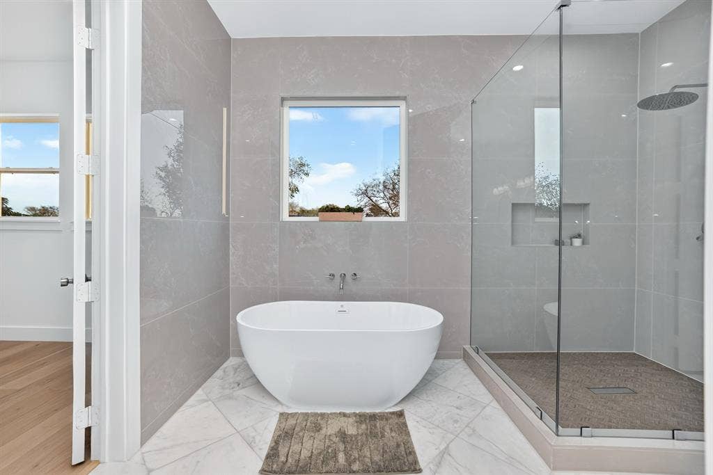 Freestanding soaking tub, walk-in glass-enclosed shower with rain-style showerhead, large-format wall tiling, recessed shower niche, and tile flooring
