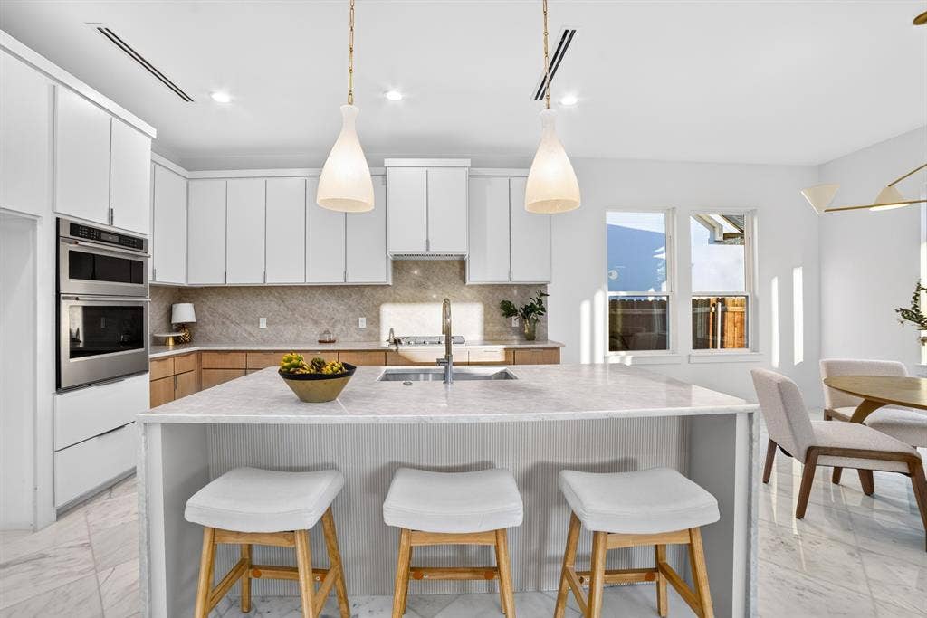 Kitchen featuring an oversized central island with stone-surface countertop, white cabinetry with wood-finish lower cupboards, stone-tiled backsplash, stainless steel double wall ovens, and pendant lighting