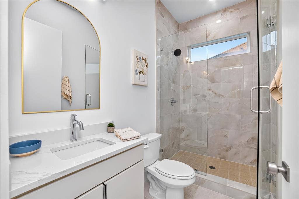 Bathroom featuring a vanity with white countertop, arched-top gold-framed mirror, and a walk-in shower with a frameless glass door and stone-finish wall tile