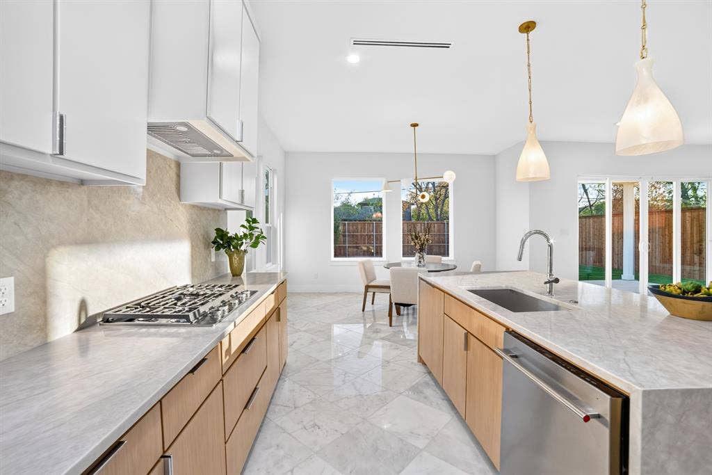 Kitchen featuring light wood-finish cabinetry, stone-patterned countertops with a matching backsplash, a large central island with an integrated sink, polished stone-tile flooring, and glass pendant lighting