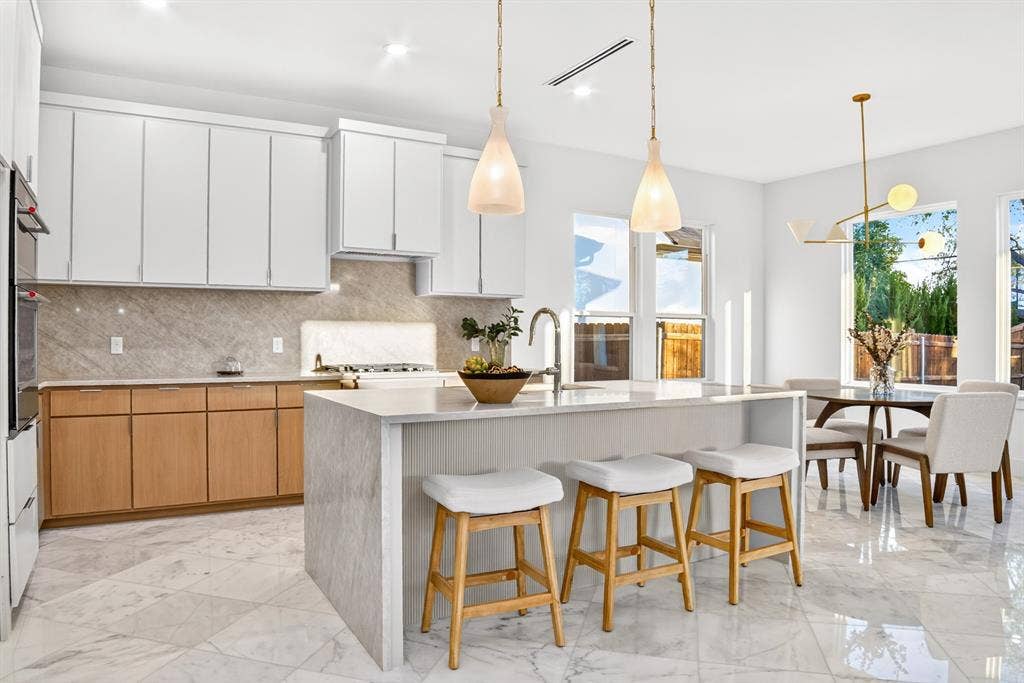 Kitchen featuring two-tone cabinetry, stone-finish backsplash, central island with waterfall-edge countertop, stone-finish flooring, and multi-light pendant fixtures