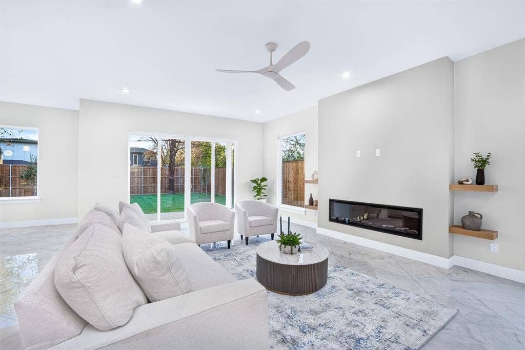 Living area featuring large-format tile flooring, a horizontal linear fireplace, wood-finish floating shelving, multi-panel sliding glass doors, and a modern ceiling fan