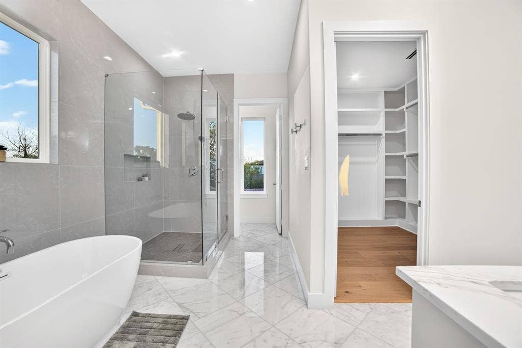 Freestanding soaking tub, glass-enclosed shower with tiled walls and recessed shelving, stone-finish tile flooring, and walk-in closet featuring built-in shelving and wood-finish flooring