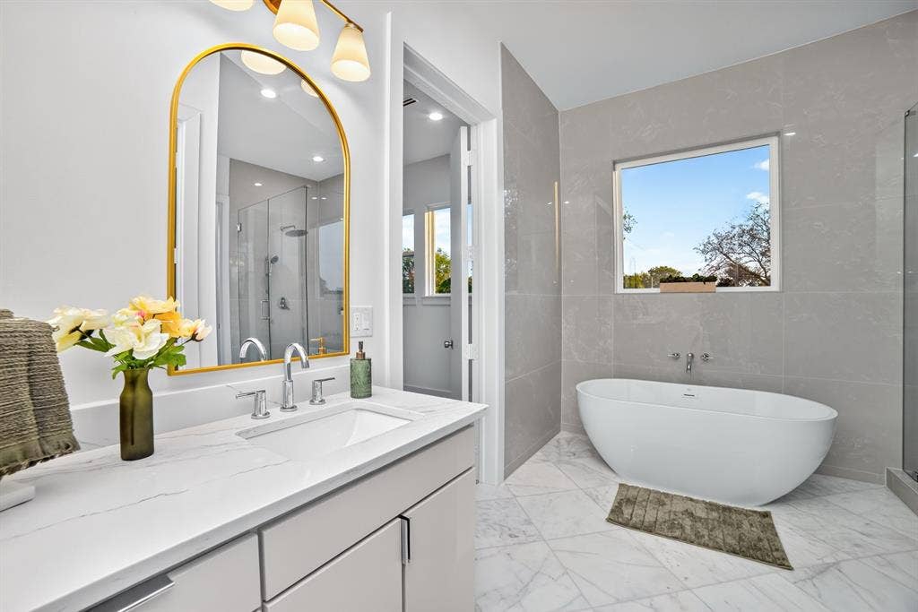 Bathroom featuring a freestanding soaking tub, arched gold-framed mirror, vanity with a white countertop and undermount sink, and large-format wall tiling