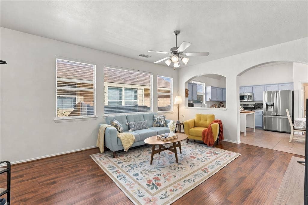 Wood-finish flooring throughout, with multiple windows, a ceiling fan with light fixture, and arched doorways leading to an adjoining kitchen with light-colored cabinetry and stainless steel appliances