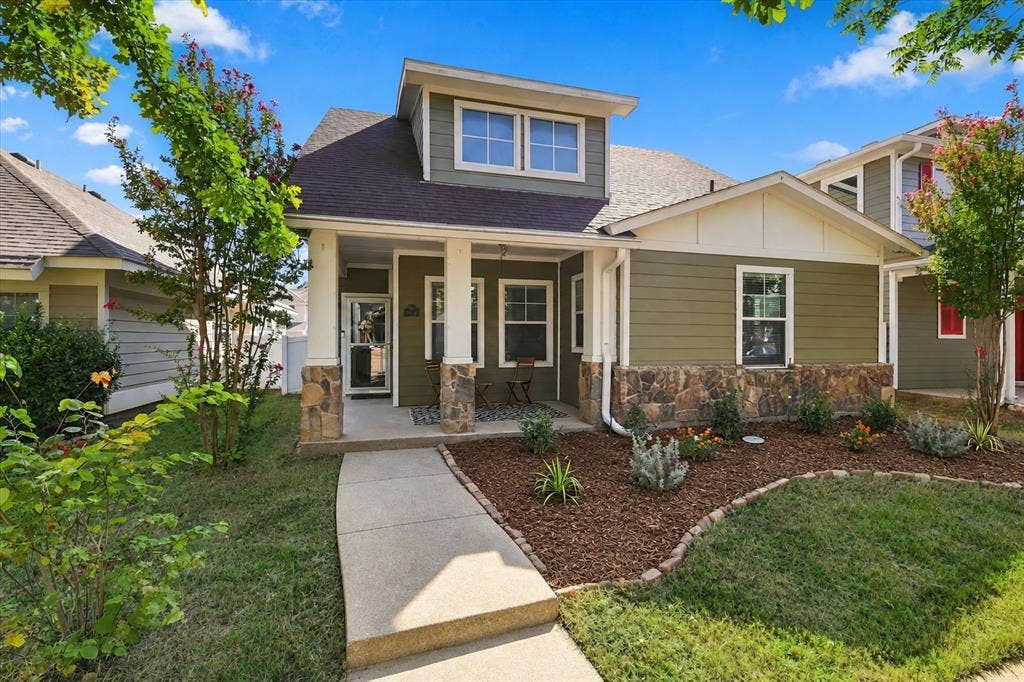 Front exterior featuring an olive green facade, stone veneer accents, an inviting covered porch, a dormer window, and a landscaped garden bed