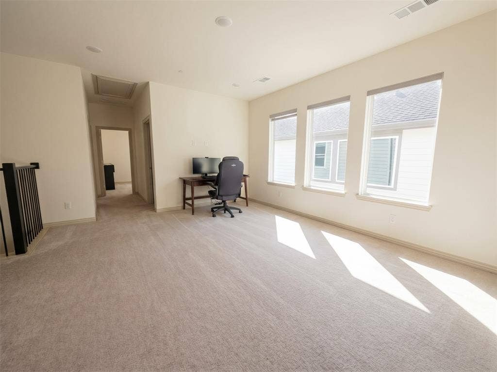 Open floor area featuring neutral wall-to-wall carpeting, recessed lighting, and three oversized windows with roller shades, accented by a black metal stair railing