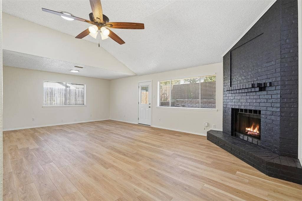 Vaulted ceiling with a ceiling fan, light wood-finish flooring, black brick fireplace, multiple windows with blinds, and a white exterior door