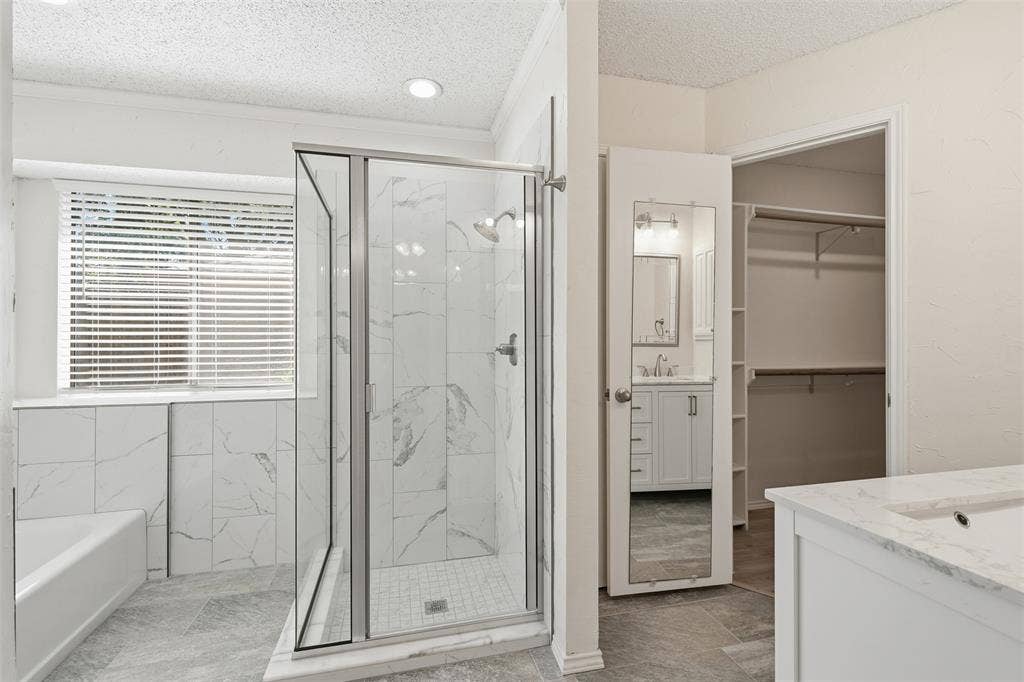 Walk-in shower with clear glass enclosure and subway tile surround, white bathtub, window with horizontal blinds, gray tile flooring, and a vanity with a white countertop