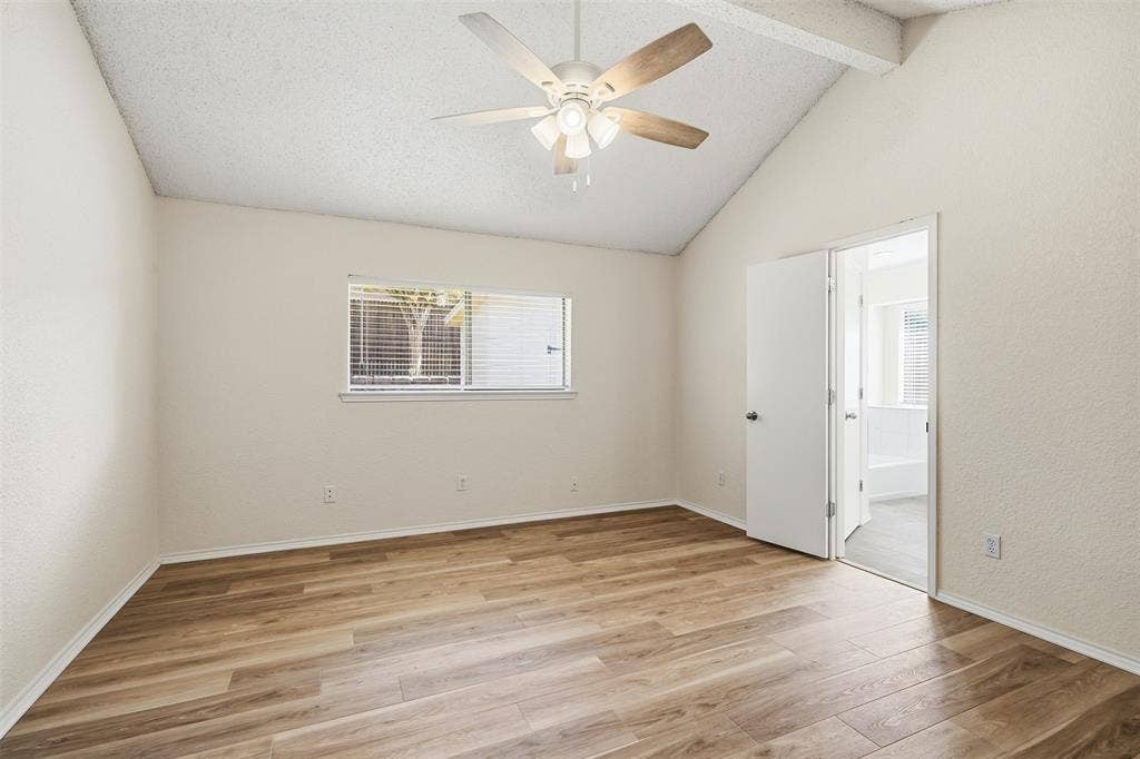 Vaulted ceiling with exposed beam, ceiling fan with light fixture, wood-finish flooring, white horizontal blinds, and a white interior door