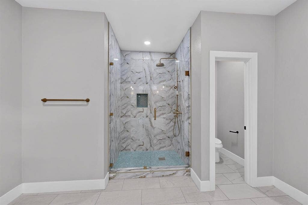 Walk-in shower featuring large-format patterned wall tile, a glass enclosure, a recessed niche, and a metallic finish shower system with a handheld sprayer