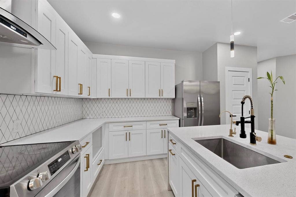 Kitchen featuring white cabinetry with gold-tone hardware, stone countertops, diamond-patterned tile backsplash, stainless steel appliances, and a central island with an undermount sink and matte black faucet