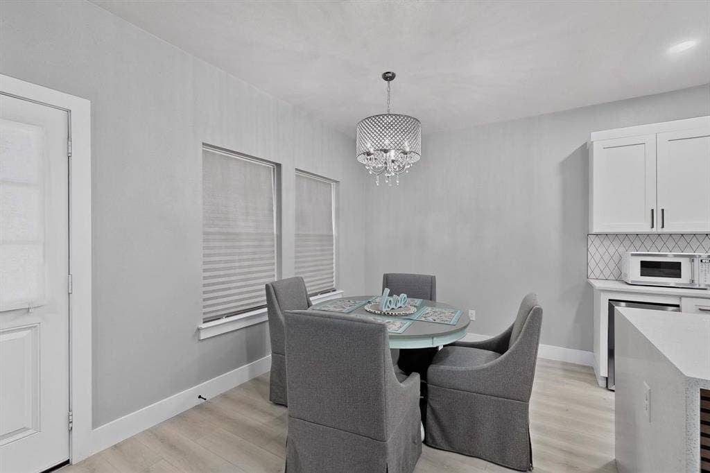 Dining area featuring wood-finish flooring, white cabinetry, a tiled backsplash, dual windows with blinds, and a crystal drum chandelier