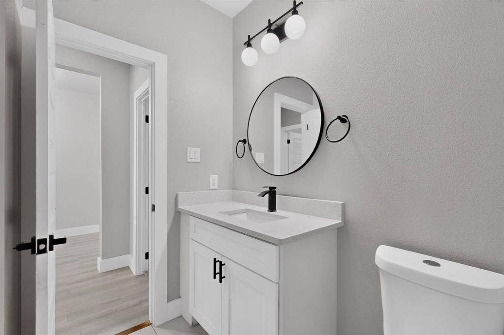 White shaker-style vanity with a solid surface countertop, matte black faucet, round mirror, and three-light vanity fixture
