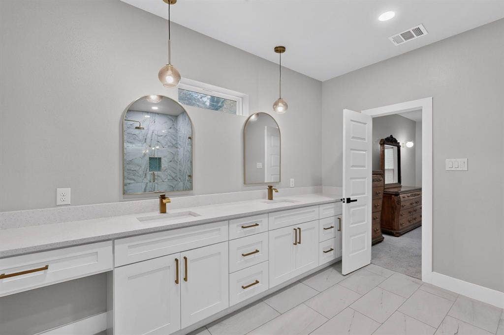 Dual-sink vanity featuring white cabinetry, brass hardware, quartz countertops, arched mirrors, and pendant lighting, finished with large-format floor tiling