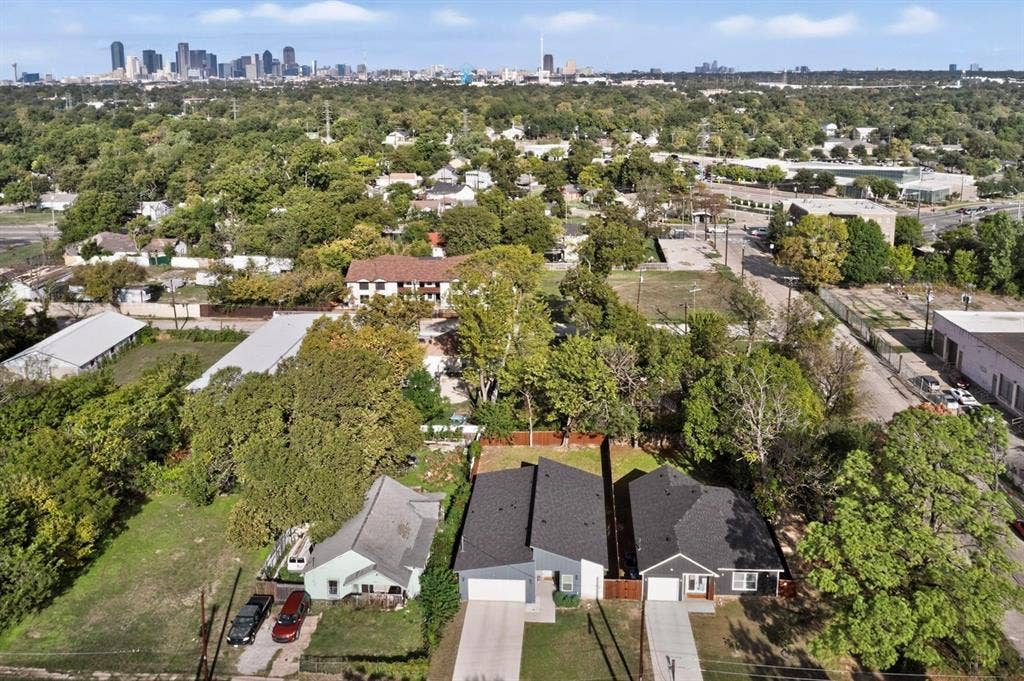 Aerial perspective showcasing residential lots with concrete driveways, attached garages, composition shingle roofs, and a distant city skyline backdrop