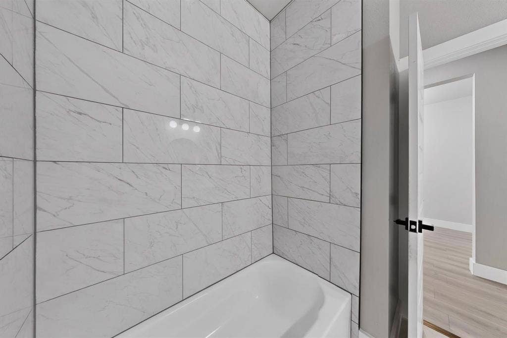 Bathtub enclosure featuring white stone-patterned tile, white acrylic soaking tub, and black door hardware