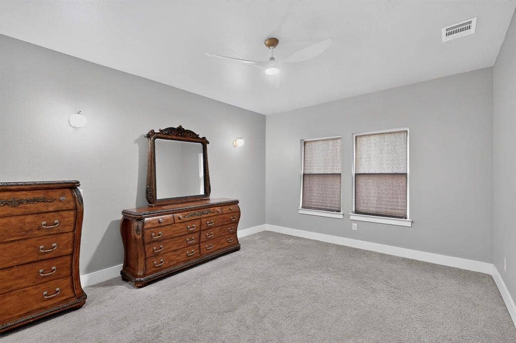 Neutral-toned interior space featuring wall-to-wall carpeting, dual windows, a ceiling fan with integrated lighting, and two wall-mounted light fixtures