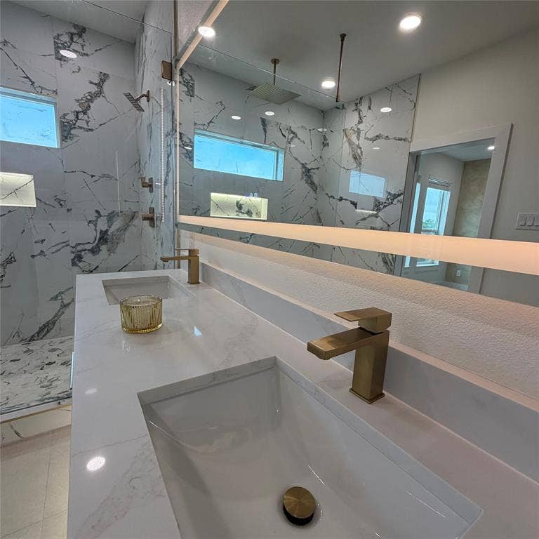Dual-sink vanity with white stone-finish countertop, brushed gold-tone faucets, integrated LED mirror, and wall-to-wall large-format porcelain tile surrounding the walk-in shower